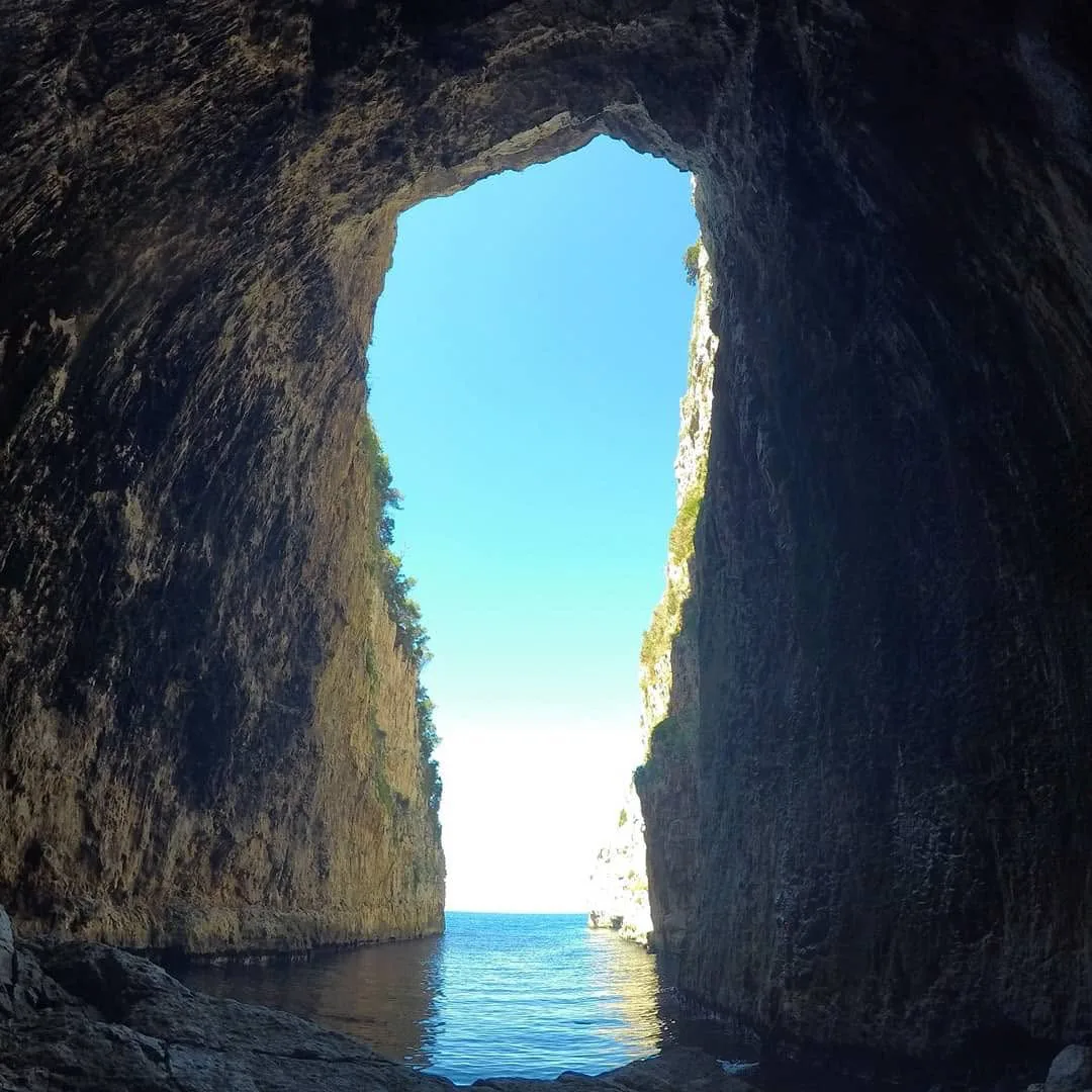 View from inside Haxhi Ali Cave through its tall arched opening to calm sea and blue sky