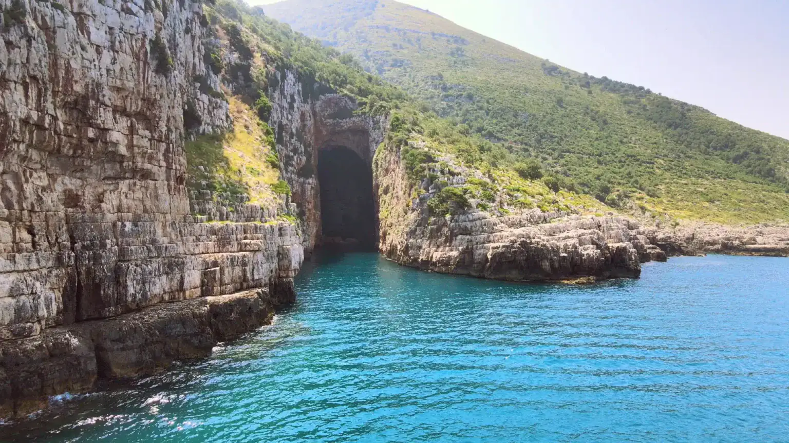 The tall dark mouth of Haxhi Ali Cave in a layered limestone cliff, with turquoise water in front and a green slope above