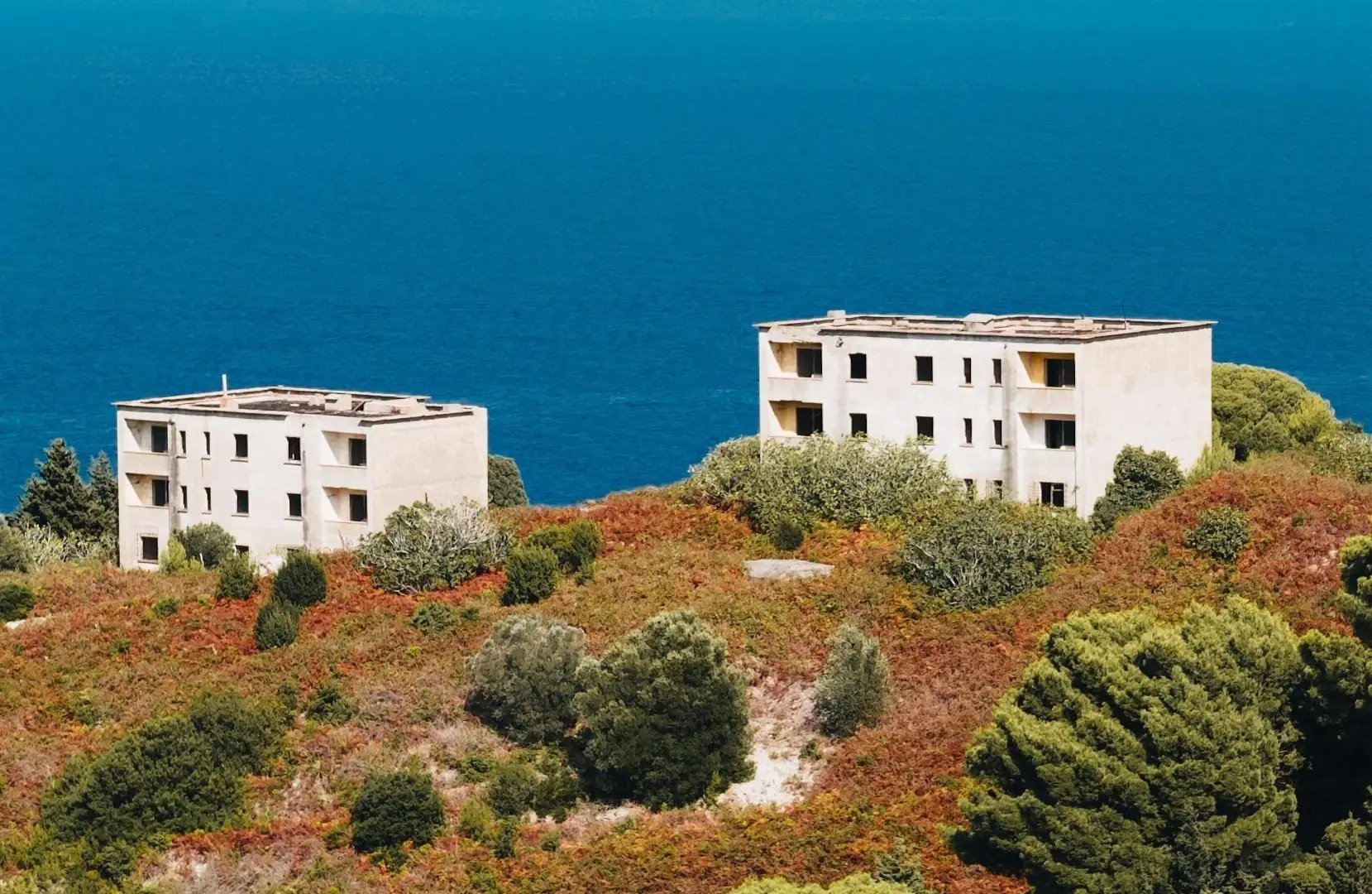 Two abandoned white multi-storey blocks on a slope of red-brown scrub and pines, with the open sea behind