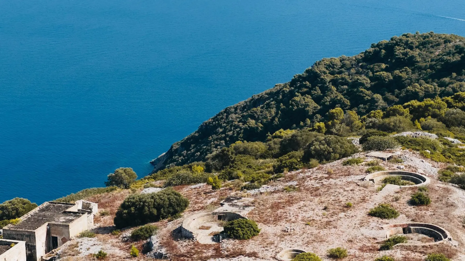 Aerial view of a dry ridge with circular concrete emplacements and a low concrete building above deep-blue sea