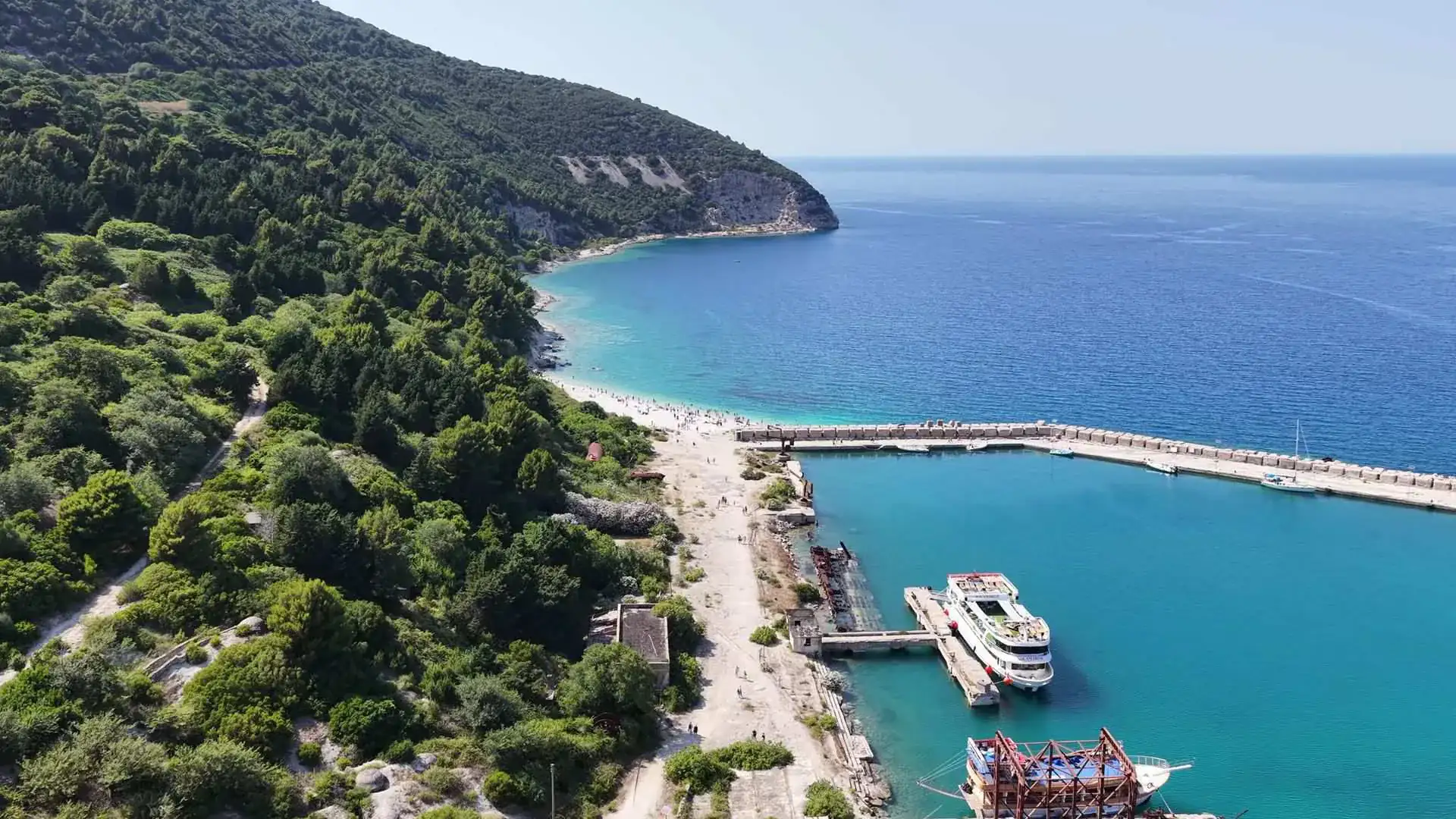 Aerial view of Sazan's landing area: a long breakwater, passenger ships at the pier, a pebble beach and pine-covered slopes