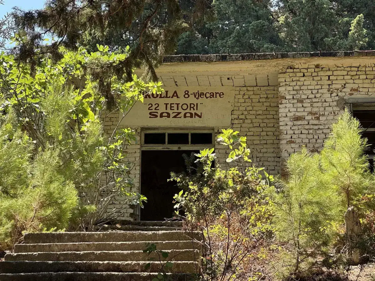 The overgrown entrance of a derelict brick building whose sign reads "Shkolla 8-vjeçare 22 Tetori Sazan"