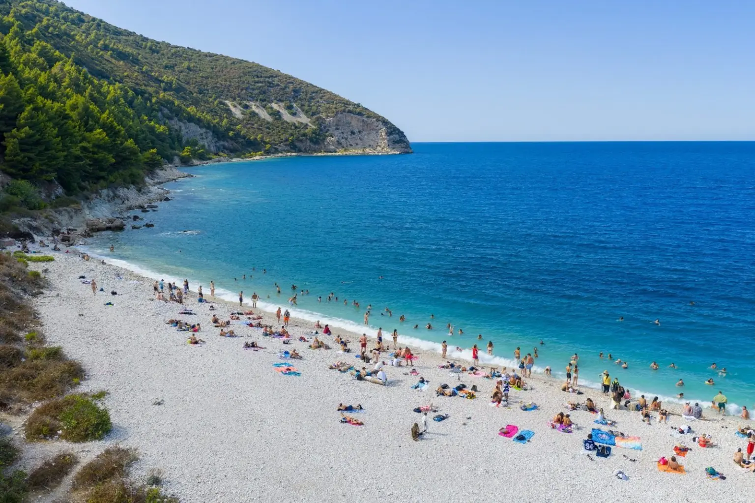 Aerial view of a long pale pebble beach on Sazan Island, with swimmers, turquoise shallows and a pine-covered headland